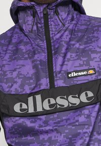 Lila vindjacka med ett digitalt kamouflage-mönster, med ett svart horisontellt rand med "ellesse"-logotyp i grått och en dragkedja framtill.