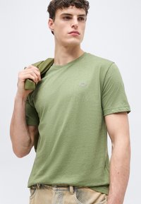 Νέος άνδρας που φοράει μια ανοιχτοπράσινη μπλούζα, κρατώντας ένα ταιριαστό σακάκι στον ώμο του, κοιτάζει στο πλάι με φόντο ένα απλό φόντο.