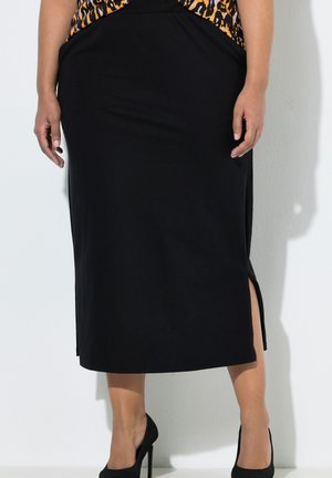 Kobieta ubrana w czarną spódnicę midi z bocznym rozcięciem, bluzkę w panterkę i czarne buty na wysokim obcasie, stoi na tle białej ściany.