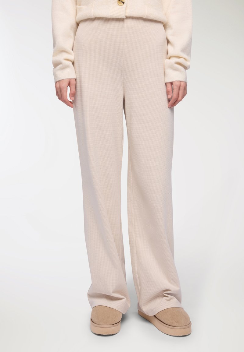 Beżowe spodnie o szerokich nogawkach z fakturowanego materiału dzianinowego, charakteryzujące się luźnym krojem o gładkiej, miękkiej powierzchni, w połączeniu z beżowym obuwiem typu slip-on.