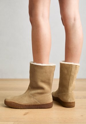Jambes portant des bottes en suède marron clair mi-mollet avec doublure en polaire crème, debout sur un sol en bois contre un mur gris clair.