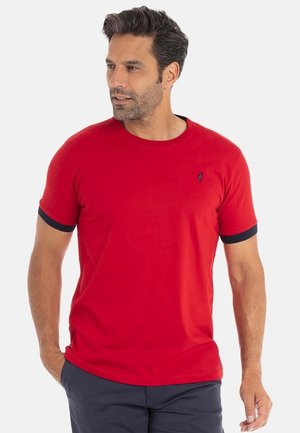 Homme portant un t-shirt rouge à manches courtes avec des bords de manches noirs et un pantalon bleu marine, regardant à gauche avec les mains dans les poches.