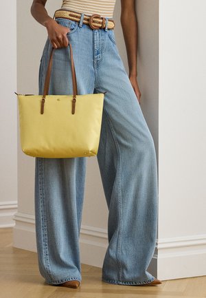 Άτομο που φοράει τζιν με φαρδιά μπατζάκια και ριγέ τοπ, κρατώντας μια κίτρινη τσάντα ώμου με καφέ δερμάτινες λαβές και πλεκτή ζώνη.