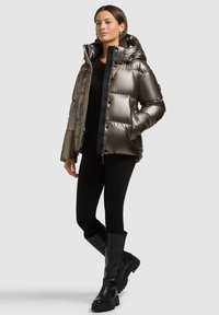 Metallic pufferjakke med hætte, trykknapper og sidelommer, parret med tætsiddende sorte bukser og knæ-høje sorte støvler.