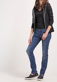 Sudadera con capucha negra con cremallera, camiseta negra ajustada, vaqueros pitillo azules y zapatillas negras sin cordones con un patrón acolchado. Atuendo casual.