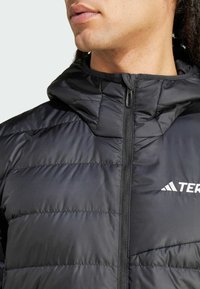Veste isolée noire avec fermeture éclair, capuche structurée et motif cousu. Le logo est affiché sur la poitrine gauche.