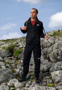 Sort sportsjakke og bukser lavet af slidstærkt stof. Har en lynlåslomme og logodetalje. Bæres med en rød indershirt og støvler.