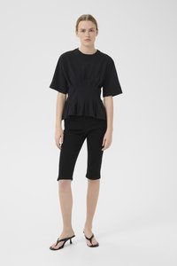 Svart kortermet topp med plissert design, sammen med svarte knelange, tettsittende shorts og svarte hælsandaler. Enkel, elegant silhuett.