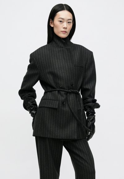 Zwarte pinstripe double-breasted blazer met hoge kraag, geweven textuur, ingesnoerd middenstuk en bijpassende wijde broek. Handschoenen voegen detail toe.