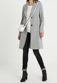 Lichtgrijze wollen jas met grote knopen, lange mouwen en een reverskraag met insnijding. Zwarte skinny jeans en enkellaarsjes. Witte schoudertas.