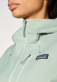 Svetlo meta zelena jakna z visokim ovratnikom, opremljena z sprednjo zadrgo, nastavljivimi vrvicami in črno oznako logotipa Patagonia.