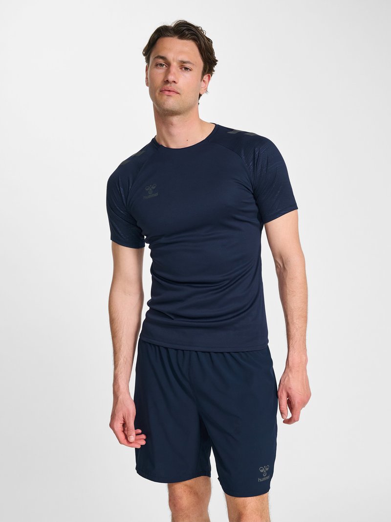 Jonge man die een marineblauw sportshirt met korte mouwen en bijpassende korte broek draagt, staand tegen een effen lichte achtergrond.