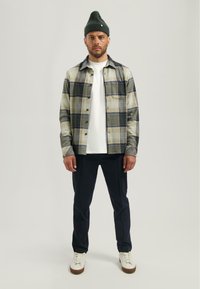 Geruit shirt in beige en groene tinten, wit T-shirt, marineblauwe broek, witte sneakers en een groene gebreide beanie. Staande tegen een neutrale achtergrond.