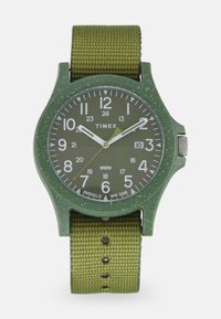 Timex-Uhr mit einem grünen Kunststoffgehäuse, weißen Ziffern und einem strukturierten grünen Nylonarmband. Verfügt über Indiglo-Beleuchtung und eine Wasserfestigkeit von 30 m.