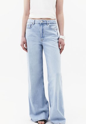 Frau trägt hellblaue Jeans mit weitem Bein und ein weißes bauchfreies Tanktop, steht mit locker an den Seiten hängenden Händen.