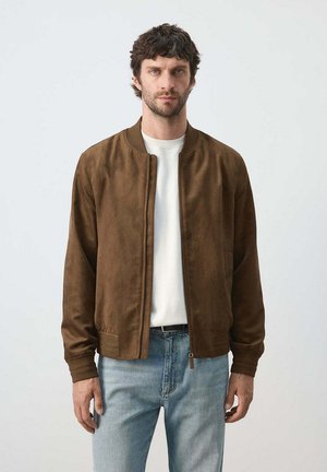 Hombre con cabello corto y rizado, lleva una chaqueta de ante marrón, camisa blanca y vaqueros azul claro, de pie frente a un fondo liso.