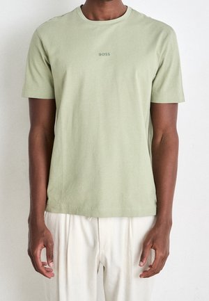Mann trägt ein hellgrünes BOSS T-Shirt mit kurzen Ärmeln und weiße Hosen, steht vor einem einfarbigen, hellen Hintergrund.