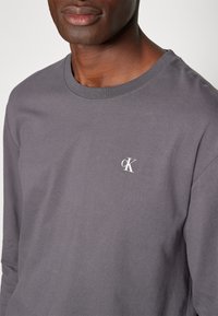 Harmaa pitkähihaine t-paita, joka on valmistettu puuvillasta. Siinä on pyöreä kaula-aukko ja pieni valkoinen logo "cK" vasemmassa rintakuvassa. Pehmeä pinta.