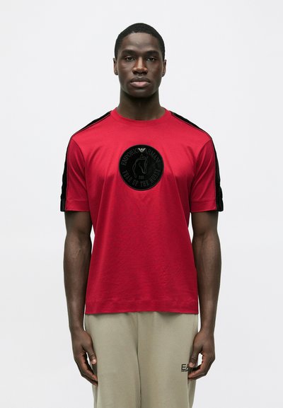 Red cotton t-shirt with short sleeves, featuring black shoulder accents and a circular black logo with a horse design on the front.