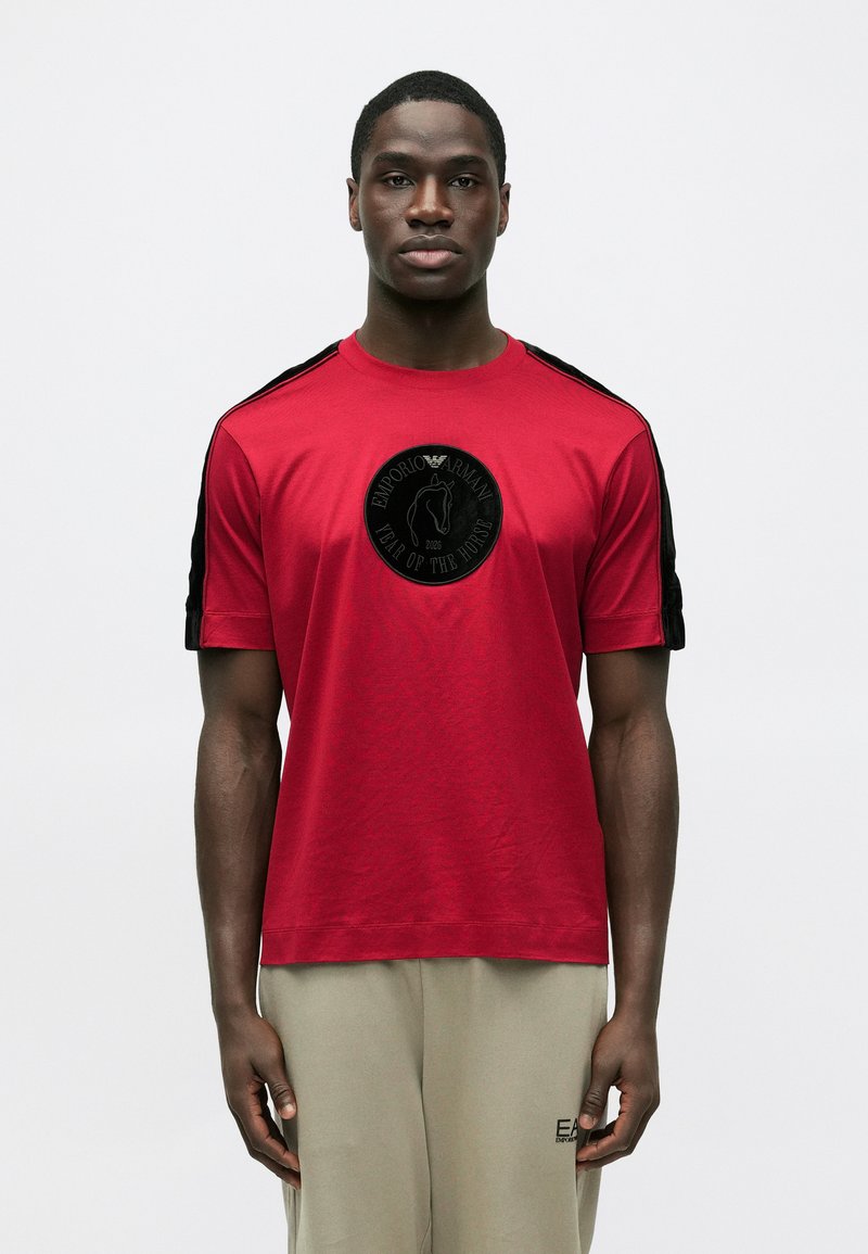 Rdeča bombažna majica s kratkimi rokavi, črnimi poudarki na ramenih in okroglim črnim logotipom s konjsko motivo spredaj.