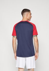 Korte mouwen sportshirt met een marineblauw lijf en rode raglanmouwen. Gemaakt van een gladde stof; heeft een ronde halslijn en een ontspannen pasvorm.