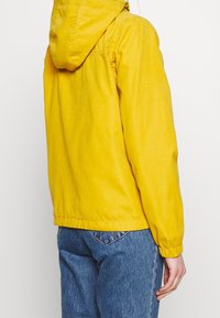 Żółta kurtka z kapturem z teksturowaną powierzchnią, elastycznymi mankietami i swobodnym krojem, zestawiona z niebieskimi dżinsami dla kontrastu.