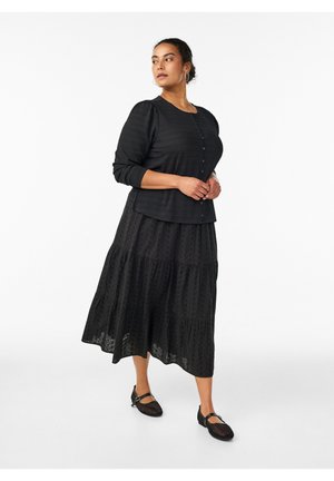 Kvinde står iført lang sort knappet bluse, matchende lagdelt nederdel og sorte Mary Jane-sko, ser til siden mod hvid baggrund.