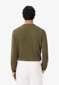 Hombre con cabello rizado oscuro, vestido con un suéter de manga larga color verde oliva y pantalones blancos, de espaldas al espectador contra un fondo blanco.
