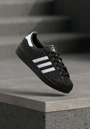 Zwarte Adidas sneaker met witte strepen, verhoogde stippen aan de zijkanten, zwarte veters en een schalenneus, zwevend boven een betonnen richel met een wazige trap op de achtergrond.