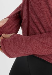 Maroon zip-up jacket with long sleeves and thumbholes, featuring a textured fabric. Close-up of hands pulling the sleeve.