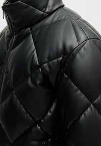 Giacca a piumino nera trapuntata realizzata in morbida e lucida ecopelle. Caratterizzata da un colletto alto e cuciture diagonali con chiusura a zip.