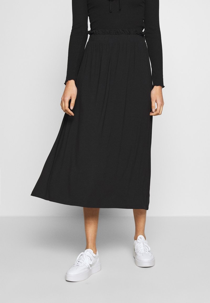 Falda negra de largo midi, hecha de una tela suave con cintura fruncida. Combinada con zapatillas blancas, de pie sobre un fondo neutro.