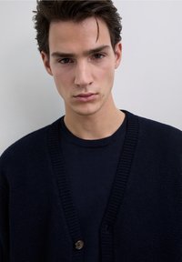 Dark navy cardigan with a V-neck and a single wooden button, worn over a matching dark top. Knit texture with ribbed edges.