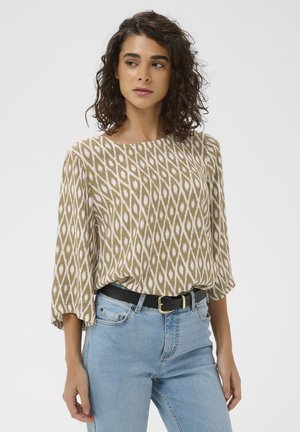 Femme aux cheveux bouclés portant un chemisier beige et blanc à motifs avec de larges manches, rentré dans un jean bleu clair avec une ceinture noire.