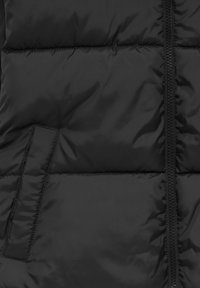 Tissu noir matelassé avec des surpiqûres horizontales, une fermeture éclair visible à droite et une ouverture de poche rectangulaire sur le côté gauche.