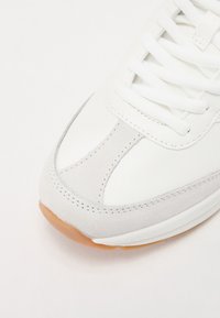 Gros plan de la partie avant d'une chaussure de sport blanche avec des détails en daim gris clair et des lacets blancs sur fond blanc.