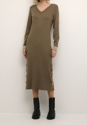 Nainen pukeutuneena pitkähihainen oliivinvihreä ribbineulottu midimekko, jossa on sivunapit, ja mustat nilkkurit, seisomassa yksinkertaisen taustan edessä.