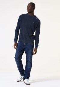Mann in navyblauer Sweatshirt, dunkelblauen Jeans und weißen Sneakers, steht und schaut zur Seite vor einem schlichten Hintergrund.
