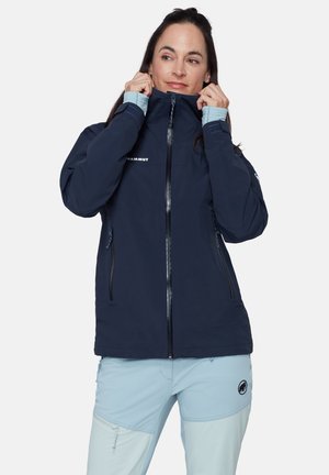 Vrouw die een marineblauw jack met Mammut-logo dichtritst, draagt lichtblauwe outdoorbroek met ritssluitingen in de zakken en Mammut-logo op de dij.