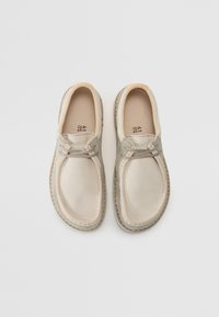 Light beige slip-on shoes with dark beige accents, soft suede texture, rounded toe, and visible stitching along the sole.