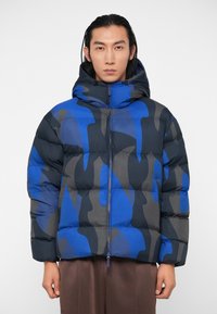 Geacă puffer în nuanțe de albastru și gri, cu un model de camuflaj, având glugă, închidere cu fermoar și textură matlasată pentru izolație.