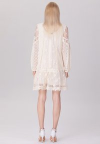 Robe en dentelle crème clair avec de longues manches volumineuses et des motifs floraux. Présente un ourlet volanté et des détails de finition délicats.