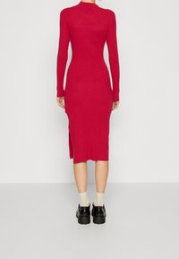 Robe rouge en maille à manches longues, ajustée, avec un décolleté rond, une fente latérale et une texture côtelée. Portée avec des chaussures noires et des chaussettes beiges.