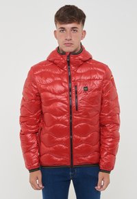 Giacca a vento rossa con cappuccio, caratterizzata da un motivo trapuntato, chiusura con zip e una piccola tasca con zip sul petto. Texture liscia e lucida.