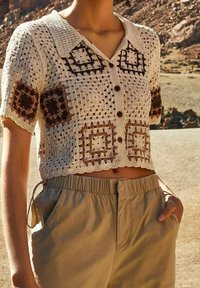 Žena v krátkém rukávu, zkráceném, knoflíkovém háčkovaném topu s hnědými a béžovými čtvercovými vzory, zkombinovaném s béžovými kalhotami s vysokým pasem.