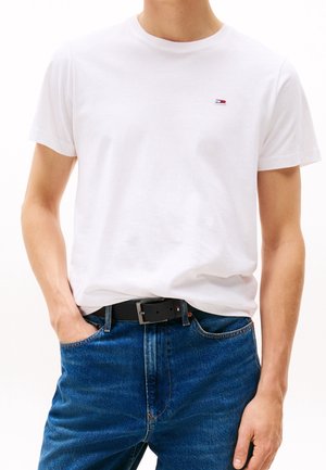 Person trägt ein weißes T-Shirt mit Rundhalsausschnitt und kleinem Logo auf der Brust, blaue Jeans und schwarzen Gürtel, steht mit einer Hand in der Tasche.