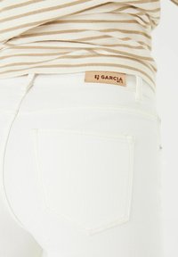 Vaqueros de mezclilla blancos con una etiqueta de marca de cuero en la cinturilla. Presenta dos bolsillos traseros y un diseño ajustado.