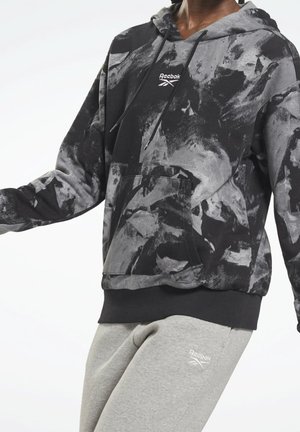 Personne portant un sweat à capuche Reebok à motifs abstraits noirs et gris et un pantalon de survêtement Reebok gris clair, posant devant un fond blanc.