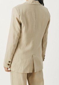 Blazer de lino beige con abertura en la espalda y puños con botones, llevado con pantalones a juego, visto desde atrás en una persona con cabello oscuro.