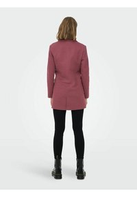 Long manteau rose avec une coupe ajustée, fente unique à l'arrière, porté par-dessus un pantalon noir et des bottes noires épaisses. Texture lisse, forme structurée.